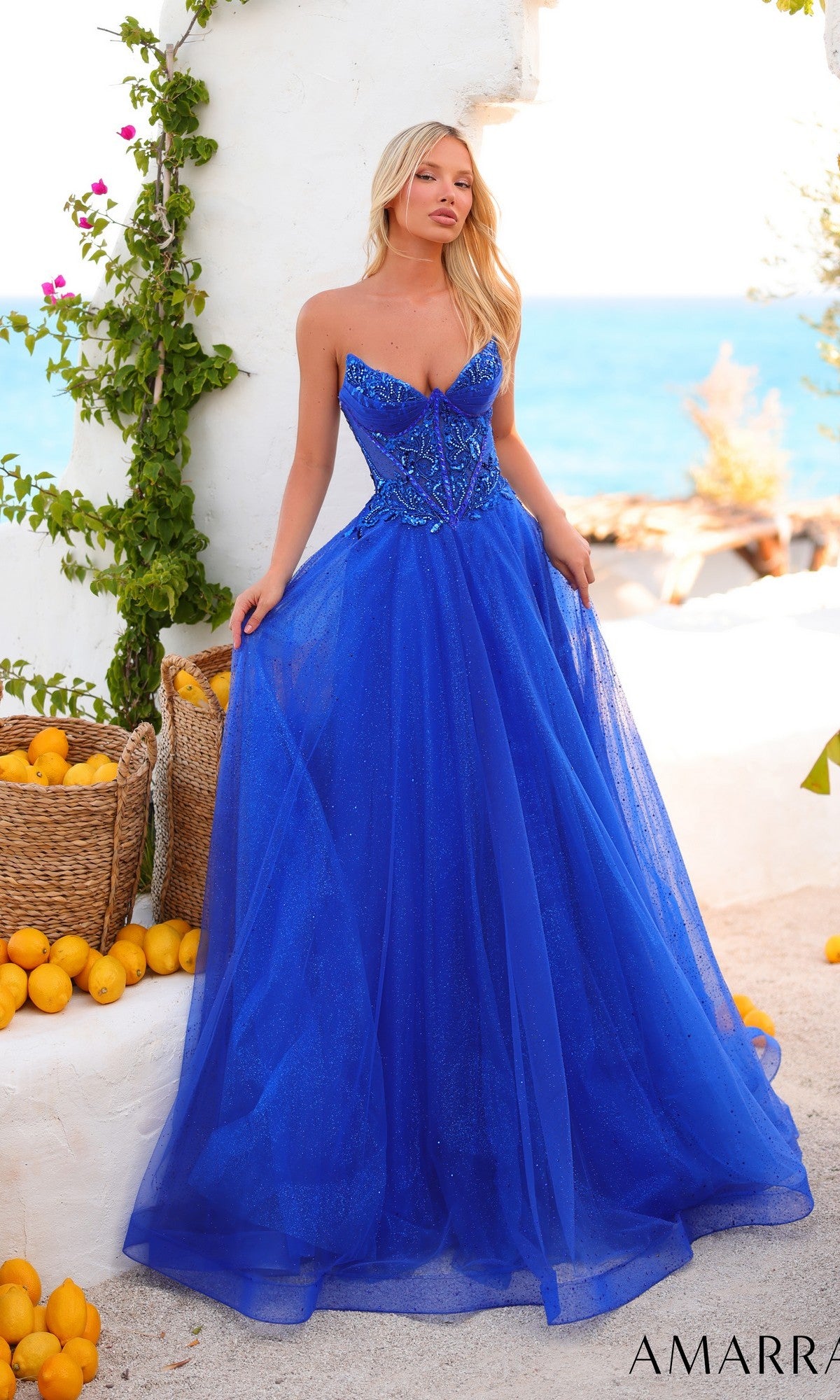 Wearing the Long Prom Dress: Amarra 89488, featuring a strapless embellished corset bodice and glitter tulle A-line skirt, a woman stands outdoors by a white wall, baskets of lemons, lush greenery, and the shimmering sea in the background.